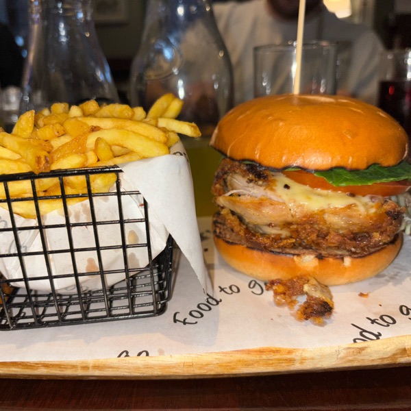 A burger in a glazed bun beside a wire basket of chips on a wooden board